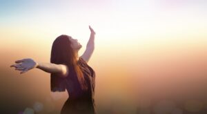 Eine Person steht mit ausgebreiteten Armen und geschlossenem Gesicht nach oben gerichtet. Die Szene ist in warmes, weiches Licht getaucht, m&ouml;glicherweise w&auml;hrend des Sonnenaufgangs oder Sonnenuntergangs.