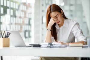 Eine Frau in wei&szlig;em Hemd sitzt gestresst an einem Schreibtisch, h&auml;lt ihre Stirn. Vor ihr liegen ein Laptop, eine Stiftbox und Papierstapel. Der Raum ist modern und hell gestaltet.