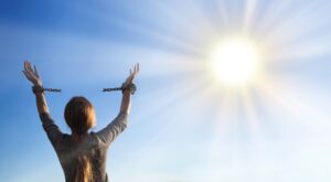 Eine Person mit offenen Armen und zerbrochenen Ketten an den Handgelenken steht drau&szlig;en unter einem klaren blauen Himmel. Die strahlende Sonne im Hintergrund symbolisiert Freiheit.