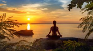 Eine Person sitzt meditierend am Rand eines Felsens am Meer, w&auml;hrend die Sonne in einem orangefarbenen Himmel untergeht. Umgeben von Bl&auml;ttern und ruhigem Wasser im Hintergrund.