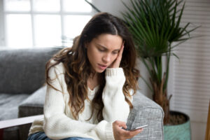 Eine Frau sitzt nachdenklich mit einem Smartphone in der Hand auf einem grauen Sofa. Im Hintergrund befindet sich eine gro&szlig;e gr&uuml;ne Pflanze und ein Fenster mit wei&szlig;en Vorh&auml;ngen.