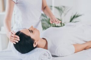 One person is lying relaxed on a table, head on a towel. Another person is gently holding their head, in a bright room with plants in the background.