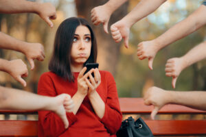 Woman in a red sweater holding a smartphone, looking worried and excited, sitting on a bench in the park, surrounded by several hands pointing thumbs down.