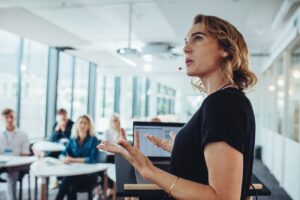 Eine Frau spricht in ein Headset-Mikrofon, w&auml;hrend sie vor Laptop-Bildschirm gestikuliert. Im Hintergrund h&ouml;ren Menschen an Tischen in einem modernen, lichtdurchfluteten Konferenzraum zu.