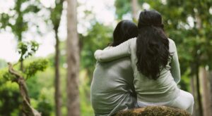 Zwei Personen sitzen eng umschlungen auf einem Felsen, tragen graue Kleidung und haben dunkles, langes Haar. Sie befinden sich in einem gr&uuml;nen Wald mit B&auml;umen und Bl&auml;ttern im Hintergrund.