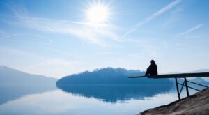 Eine Person sitzt am Ende eines Stegs, der &uuml;ber einen ruhigen See ragt. Der Himmel ist klar und sonnig, w&auml;hrend sich H&uuml;gel am Horizont erstrecken und sich im Wasser spiegeln.