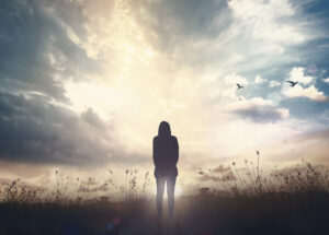 Eine Person steht still in einer Wiese und blickt in den sonnendurchfluteten Himmel. M&ouml;wen fliegen in der Ferne, w&auml;hrend Wolken dramatisch den Himmel bedecken.