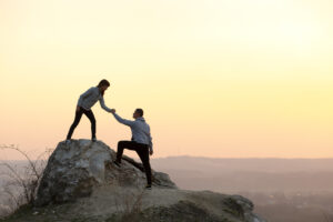 Zwei Personen interagieren auf einem Felsen bei Sonnenuntergang. Eine Person hilft der anderen, indem sie die Hand reicht. Der Hintergrund zeigt eine h&uuml;gelige Landschaft mit sanftem Licht.
