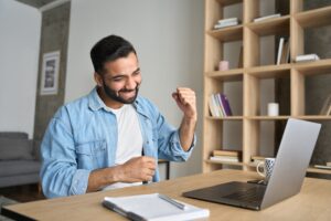 Ein Mann sitzt an einem Tisch und schaut l&auml;chelnd auf einen Laptop, w&auml;hrend er die Faust triumphierend hebt. Im Hintergrund steht ein Regal mit B&uuml;chern und Dekoration.