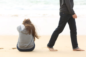 Eine Person sitzt mit gesenktem Kopf am Strand, w&auml;hrend eine andere Person barfu&szlig; vorbeigeht. Im Hintergrund sind Wellen des Meeres zu sehen, die eine ruhige, nachdenkliche Stimmung schaffen.