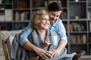 Eine &auml;ltere Frau sitzt mit einer Tasse in der Hand, eingewickelt in eine Strickdecke. Ein j&uuml;ngerer Mann umarmt sie von hinten. Im Hintergrund ist ein B&uuml;cherregal sichtbar.