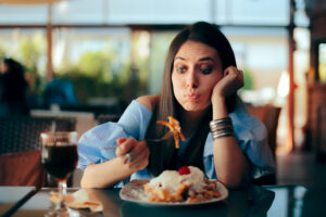 Eine Frau sitzt an einem Tisch, h&auml;lt eine Gabel mit Spaghetti in der einen Hand und st&uuml;tzt ihren Kopf in die andere Hand. Sie schaut skeptisch. Im Hintergrund ist ein Restaurant zu sehen.