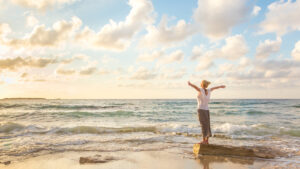 Eine Person steht mit ausgebreiteten Armen auf einem Felsen am Strand, w&auml;hrend Wellen leicht an das Ufer schlagen. Der Himmel ist von dichten, wei&szlig;en Wolken bedeckt und vermittelt eine friedliche Atmosph&auml;re.
