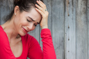 Eine Frau mit einem roten Oberteil h&auml;lt ihren Kopf mit einer Hand, scheinbar in emotionalem Schmerz, vor einem grauen Holzhintergrund. Kein Text ist im Bild enthalten.