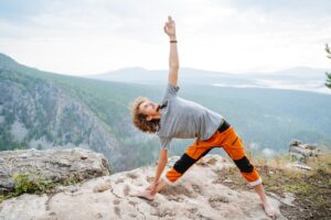 Eine Person in sportlicher Kleidung f&uuml;hrt eine Yoga-&Uuml;bung auf einem felsigen Plateau aus, umgeben von bergiger Landschaft und bew&ouml;lktem Himmel, mit weitreichendem Ausblick auf W&auml;lder und T&auml;ler.