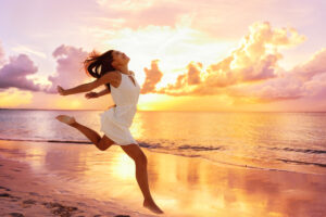 Eine Person in einem wei&szlig;en Kleid springt am Strand, beleuchtet von einem spektakul&auml;ren Sonnenuntergang. Der Himmel ist mit gelben und rosa Wolken gef&auml;rbt, das Meer reflektiert das Licht.