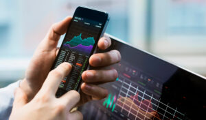 Ein Smartphone zeigt Aktienkurse und Diagramme, w&auml;hrend eine Hand den Bildschirm ber&uuml;hrt. Im Hintergrund befindet sich ein Laptop mit &auml;hnlichen Finanzdiagrammen, in einem B&uuml;ro oder Arbeitsumfeld.