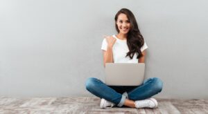 Eine Frau sitzt mit gekreuzten Beinen auf dem Boden, l&auml;chelt und zeigt mit dem Daumen zur Seite. Sie hat einen Laptop auf dem Scho&szlig;, der vor einer grauen Wand steht.
