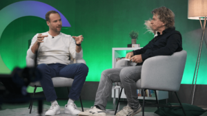 Two men sit in gray armchairs and talk in a studio with a green background. One is gesticulating animatedly while the other is taking notes. A shelf and a lamp are in the background.