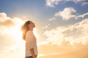 Eine Person steht seitlich im Sonnenlicht, schaut nach oben und wirkt entspannt. Der Himmel ist mit einigen wei&szlig;en Wolken bedeckt, w&auml;hrend die Sonne intensiv scheint, was eine warme Atmosph&auml;re schafft.