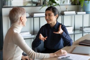 Zwei Personen sitzen an einem Tisch in einem B&uuml;ro und unterhalten sich lebhaft. Eine Person gestikuliert, w&auml;hrend die andere aufmerksam zuh&ouml;rt. Im Hintergrund sind B&uuml;cherregale mit Pflanzen und Akten sichtbar.