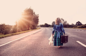 A child with long hair, holding a teddy bear and a suitcase, walks barefoot along an empty street. The scenery is rural with trees and sunset in the background.