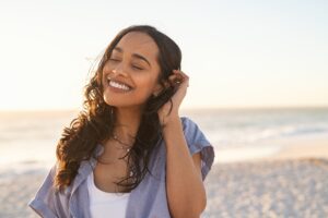 A woman smiles with her eyes closed as she holds a hand to her face. She is standing on the beach at sunset, with the sea in the background and the light of day.