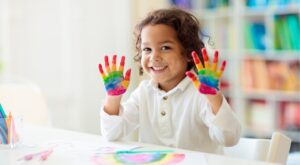 Ein Kind mit lockigem Haar zeigt l&auml;chelnd seine mit Regenbogenfarben bemalten Handfl&auml;chen. Im Hintergrund sind bunte B&uuml;cher in einem Regal sichtbar. Der Tisch ist mit Malutensilien bedeckt.