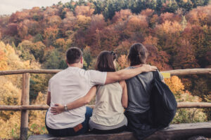 Drei Personen sitzen eng nebeneinander auf einer Holzbank, eine Person legt Arme um die anderen beiden. Sie blicken auf einen herbstlichen Wald mit buntem Laub.