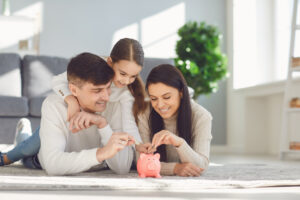 Eine Familie liegt l&auml;chelnd auf einem Wohnzimmerteppich und steckt M&uuml;nzen in ein rosafarbenes Sparschwein. Im Hintergrund steht eine gr&uuml;ne Topfpflanze neben einem Sofa und einem Fenster mit einfallendem Sonnenlicht.