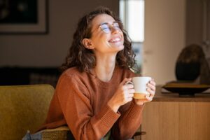 Eine l&auml;chelnde Person mit Brille h&auml;lt eine Tasse in beiden H&auml;nden. Sie tr&auml;gt einen braunen Pullover und sitzt in einem gem&uuml;tlichen Raum mit Holzm&ouml;beln im Hintergrund.