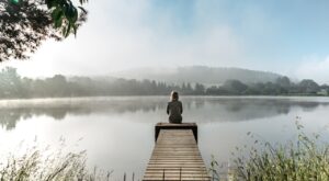 Eine Person sitzt auf einem Holzsteg und blickt auf einen ruhigen, nebligen See, der von B&auml;umen umgeben ist. Der Himmel ist leicht bew&ouml;lkt, und das Wasser spiegelt die Landschaft wider.