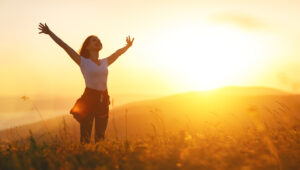 Eine Person steht mit ausgebreiteten Armen in einem sonnenbeschienenen Feld, w&auml;hrend die Sonne am Horizont aufgeht und die Umgebung in warmen Gelbt&ouml;nen beleuchtet. Gr&auml;ser bewegen sich sanft im Wind.