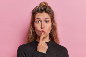 Eine Person mit br&uuml;nettem Haar ber&uuml;hrt nachdenklich ihre Lippen mit einem Finger. Sie tr&auml;gt einen dunklen Pullover. Der Hintergrund ist einfarbig rosa. Es gibt keinen Text im Bild.