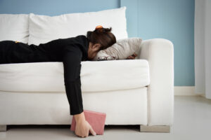 Person liegt auf einem wei&szlig;en Sofa, Gesicht im Kissen, Arm h&auml;ngt mit Buch herunter. Umgebung ist ein Raum mit hellblauer Wand und wei&szlig;em Boden, ruhig und schlicht gestaltet.