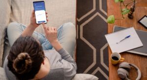 Eine Person sitzt auf einem Sofa und bedient ein Smartphone mit einem Aktienkursdiagramm. Ein Tisch daneben h&auml;lt Notizblock, Stift, Laptop, Kaffeetasse, Kopfh&ouml;rer und eine Pflanze.