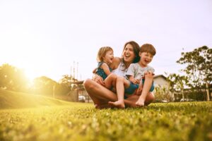 Eine Frau sitzt auf einer Grasfl&auml;che, lachend mit zwei Kindern im Arm. Die Sonne scheint hell im Hintergrund, umgeben von B&auml;umen und einem Geb&auml;ude.