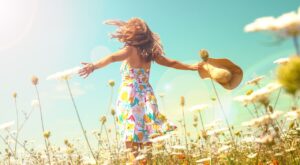 Person im bunten Sommerkleid steht mit ausgebreiteten Armen in einem bl&uuml;henden Blumenfeld, h&auml;lt einen Sonnenhut, w&auml;hrend Sonnenlicht die Szene warm beleuchtet, unter einem klaren, blauen Himmel.