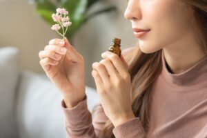Eine Person h&auml;lt einen kleinen Blumenstrau&szlig; und riecht an einer ge&ouml;ffneten Flasche mit &auml;therischem &Ouml;l. Sie sitzt in einem hellen Raum mit gr&uuml;nen Pflanzen im Hintergrund.