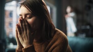 Eine Frau sitzt mit geschlossenen Augen, h&auml;lt ihre H&auml;nde vor das Gesicht und wirkt nachdenklich. Im Hintergrund ist eine weitere Person unscharf zu sehen, m&ouml;glicherweise in einem Wohnraum.