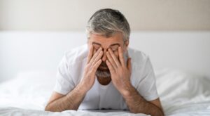 Ein Mann mit grauen Haaren reibt sich m&uuml;de die Augen. Er sitzt auf einem wei&szlig;en Bett in einem hellen, minimalistischen Raum.