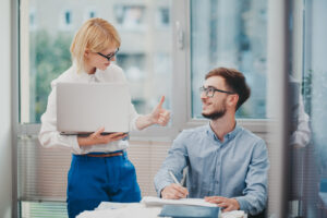 Zwei Personen interagieren in einem B&uuml;ro, w&auml;hrend eine Frau mit Laptop dem sitzenden Mann zustimmend den Daumen hebt. Sie blicken l&auml;chelnd einander an. Gro&szlig;e Fenster und Dokumente umgeben sie.