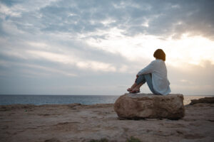 Eine Person sitzt nachdenklich auf einem gro&szlig;en Felsen am Strand und blickt aufs Meer. Die Umgebung ist ruhig, mit bew&ouml;lktem Himmel bei Sonnenuntergang.