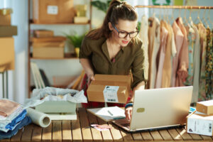 Eine Person mit Brille h&auml;lt ein Paket und arbeitet an einem Laptop in einem B&uuml;ro, umgeben von Kleidung und Verpackungsmaterialien. Im Hintergrund sind Regale mit weiteren Paketen sichtbar.