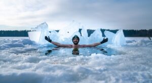 Ein Mann schwimmt in einem eisigen See, hebt die Arme und tr&auml;gt eine M&uuml;tze und Handschuhe. Im Hintergrund sind Schnee und gro&szlig;e Eisbl&ouml;cke sichtbar, umgeben von Wald.