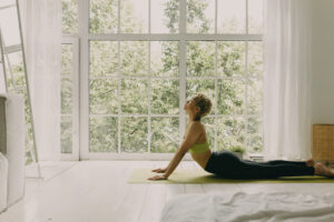 Eine Person macht eine Yoga-&Uuml;bung auf einer Matte in einem hellen Raum mit gro&szlig;en Fenstern und Blick auf gr&uuml;ne B&auml;ume. Der Raum ist minimalistisch eingerichtet und wirkt ruhig.