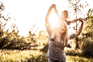 Eine Frau tr&auml;gt ein kleines Kind auf ihren Schultern, beide lachend, w&auml;hrend sie im Freien stehen. Die tiefstehende Sonne strahlt durch B&auml;ume und Gras im Hintergrund.