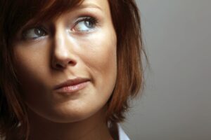 Eine Frau mit braunen Haaren schaut leicht nach rechts oben. Ihr Gesicht zeigt einen nachdenklichen Ausdruck. Der Hintergrund ist einfarbig grau, ohne erkennbare Details.