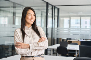 A smiling person with long dark hair stands with folded arms in a modern office with glass partitions and desks in the background.