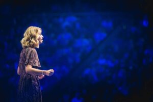 A woman stands at the side of a stage, giving a speech. In the background are blurred rows of spectators in blue. She is wearing a glittering dress and has blonde, wavy hair.